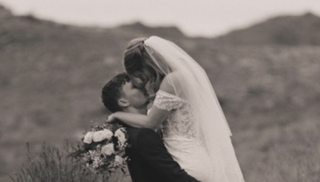 A groom lifts his bride wearing a veil and lace dress as they embrace outdoors against a hilly background.