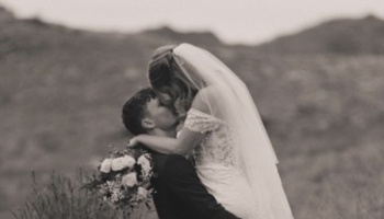 A groom lifts his bride wearing a veil and lace dress as they embrace outdoors against a hilly background.