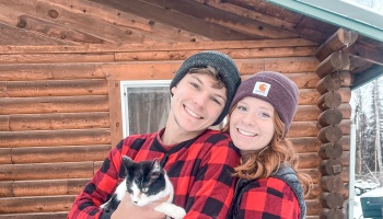 A smiling couple wearing beanies and red plaid shirts holds a black-and-white cat in front of a log cabin.