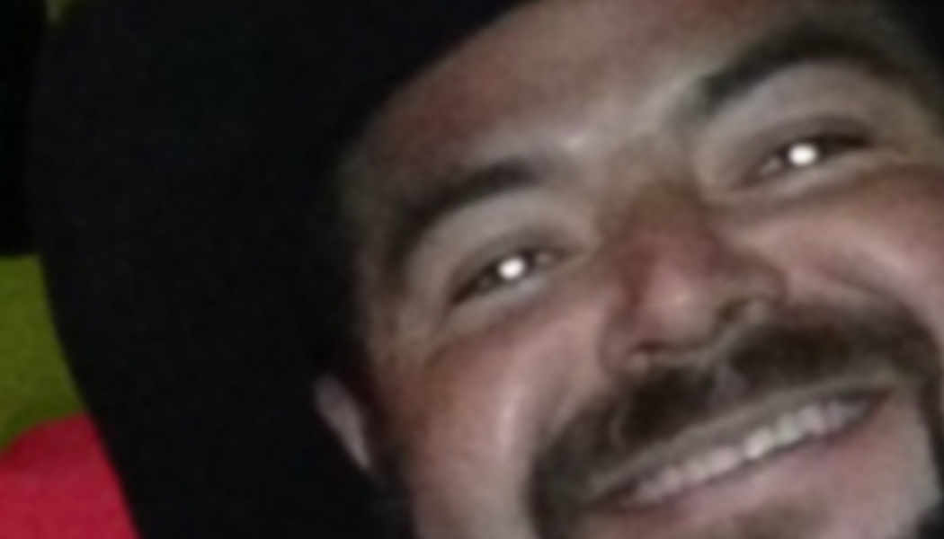 Close-up of a man with a mustache and beard smiling broadly while wearing a dark hat.