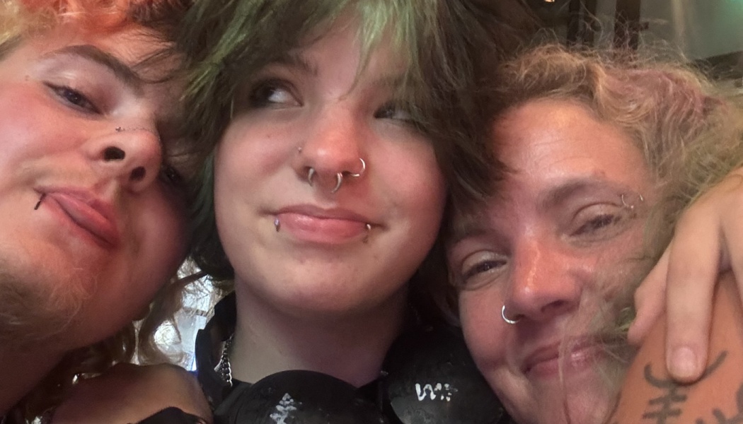 Three smiling people with facial piercings and colorful hair hugging tightly in a close-up selfie.