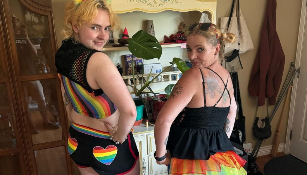 Two smiling people pose in colorful rainbow Pride outfits, looking back over their shoulders.