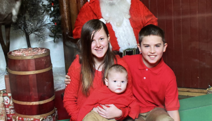 Three smiling children dressed in red pose for a Christmas photo with Santa standing behind them.