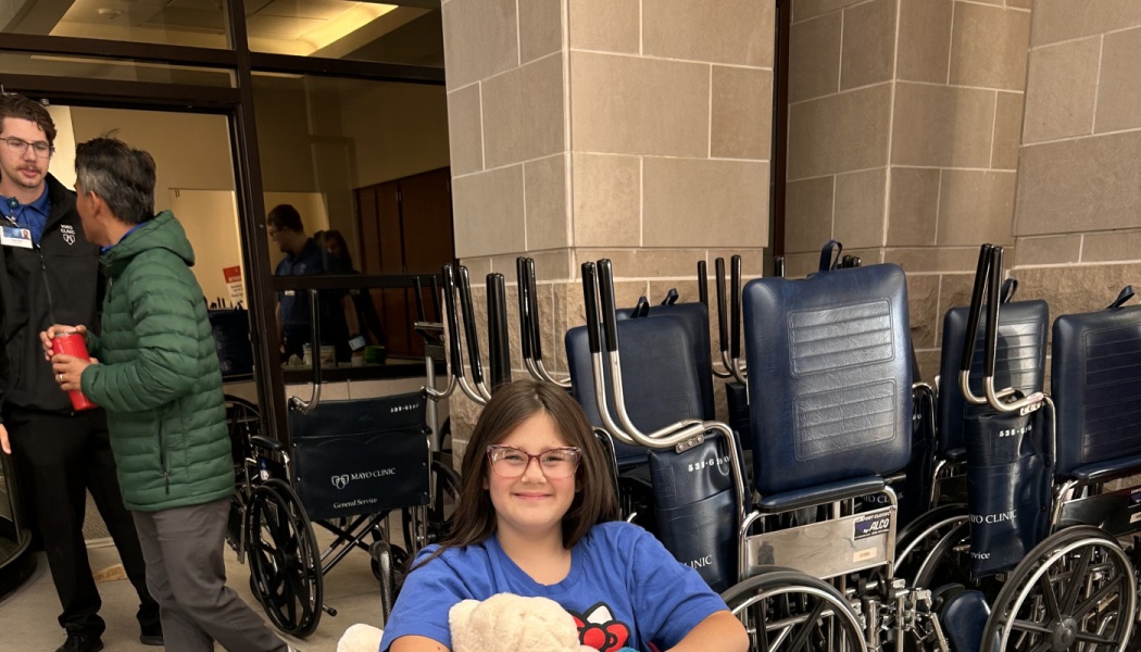 Smiling young girl with glasses sitting in a wheelchair outside Mayo Clinic, holding a stuffed animal.