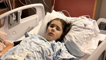 Young girl lying in a hospital bed wearing a patterned gown, connected to medical IV lines.