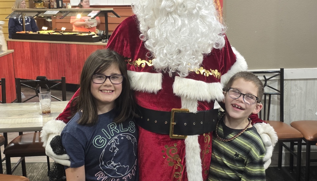 A smiling young girl and boy wearing glasses pose warmly with Santa Claus.