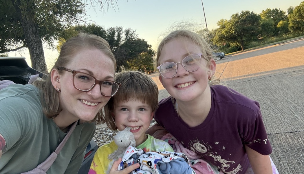A smiling woman with glasses poses outdoors with two young children, one holding a small plush toy.