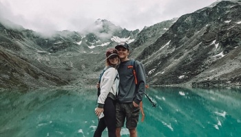 A smiling young couple poses in front of a vibrant turquoise mountain lake framed by rugged alpine peaks.