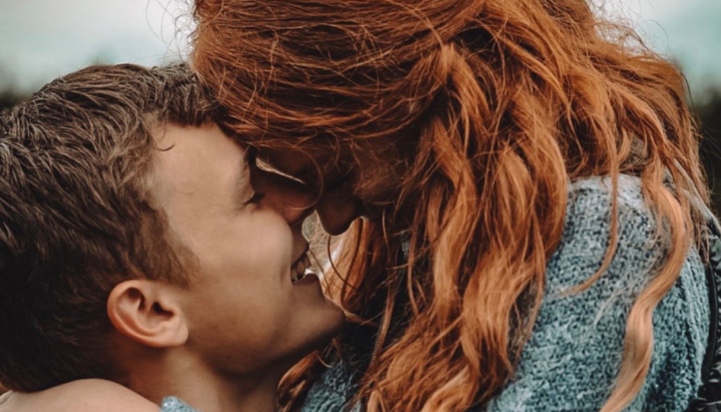 A young man smiles warmly as he touches foreheads with a red-haired woman in a tender embrace.
