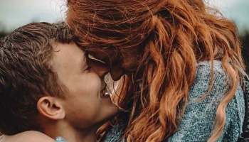 A young man smiles warmly as he touches foreheads with a red-haired woman in a tender embrace.