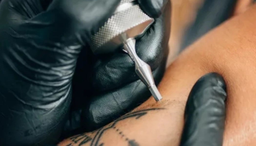 Close-up of a gloved hand holding a silver tattoo machine, applying black ink onto a person's skin.