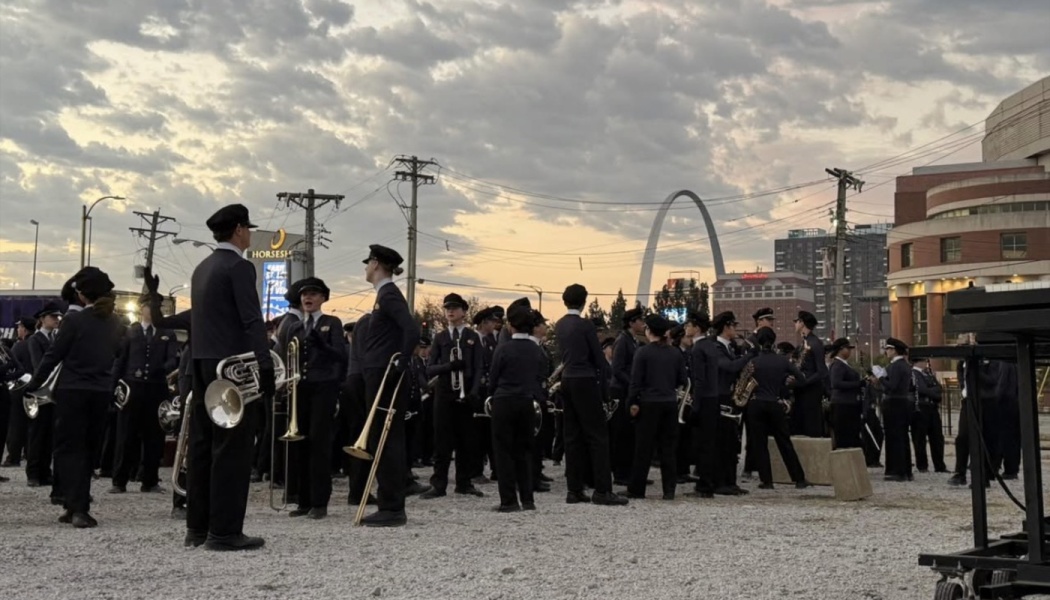 Uniformed marching band members gather outdoors with brass instruments, framed by the St. Louis Gateway Arch at dusk.