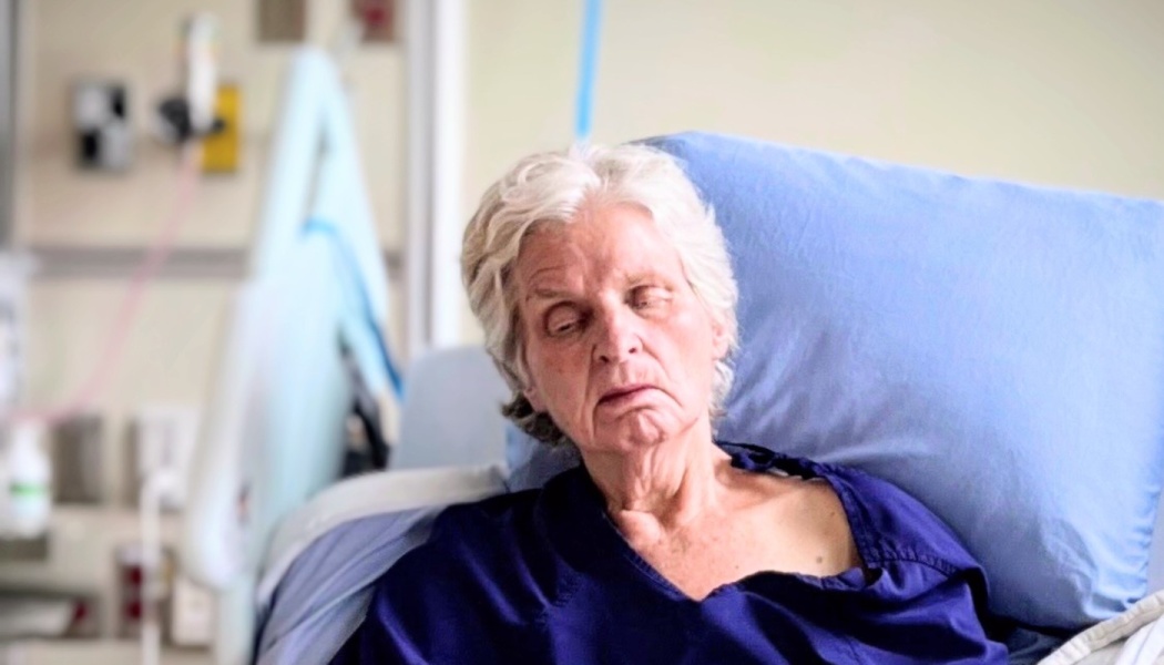 An elderly woman with short grey hair rests in a hospital bed against a blue pillow.