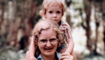A smiling woman wearing glasses carries a young toddler on her shoulders.