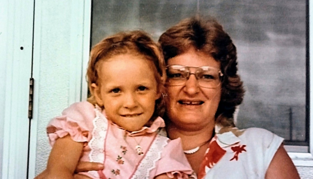 A smiling woman with glasses holds a young girl in a pink lace dress.
