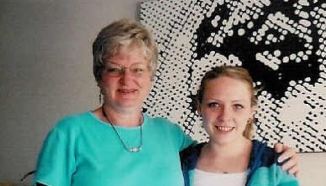 An older woman with glasses in a turquoise shirt smiles alongside a younger woman in a blue jacket.