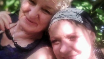 Two women smile closely together outdoors in dappled sunlight, one wearing a patterned headscarf.