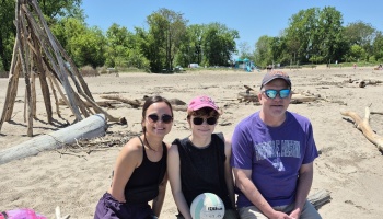 A family of three wearing sunglasses smiling together while sitting on a sandy beach on a sunny day.