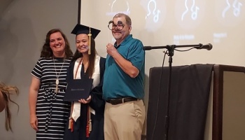 A smiling graduate holding a diploma poses with a woman and a man wearing funny novelty prop glasses.