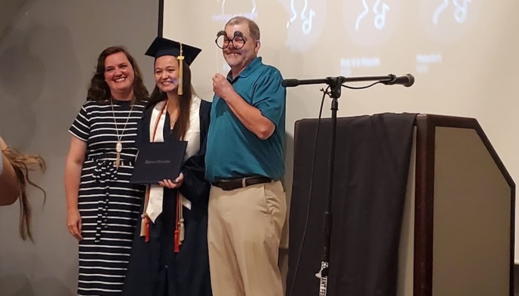 Smiling graduate in cap and gown holds diploma next to a woman and a man holding silly novelty glasses.