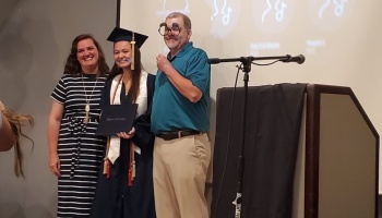 Smiling graduate in cap and gown holds diploma next to a woman and a man holding silly novelty glasses.