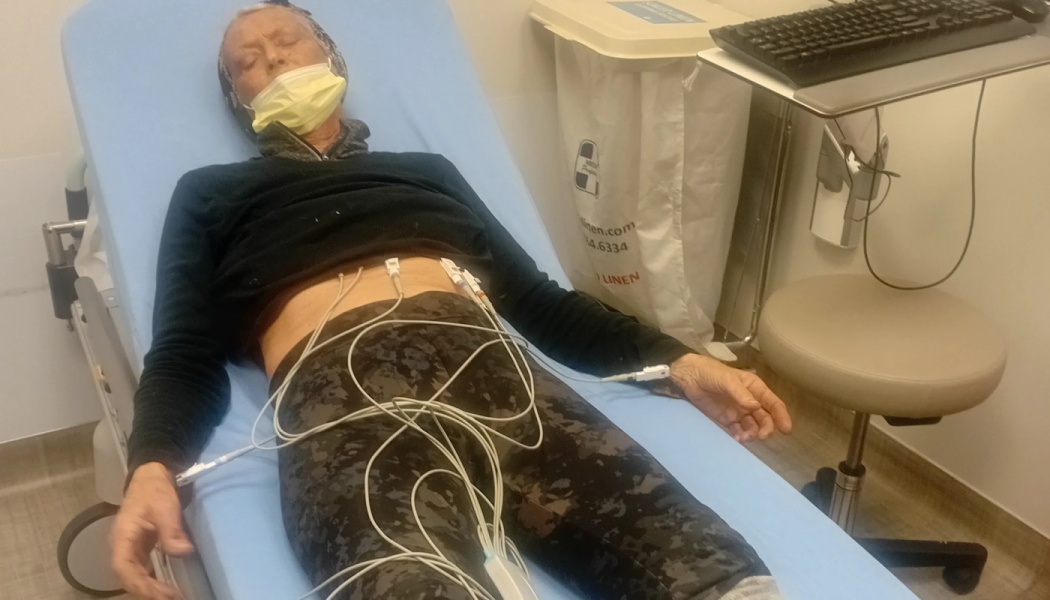 An elderly person lies on a hospital bed with ECG monitor wires attached to their abdomen.
