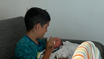 A young boy lovingly bottle-feeds a swaddled newborn baby on a couch.