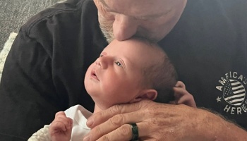 An older man tenderly holds a newborn baby and kisses the top of their head.