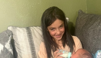 A smiling young girl sitting on a couch, feeding a newborn baby with a bottle.