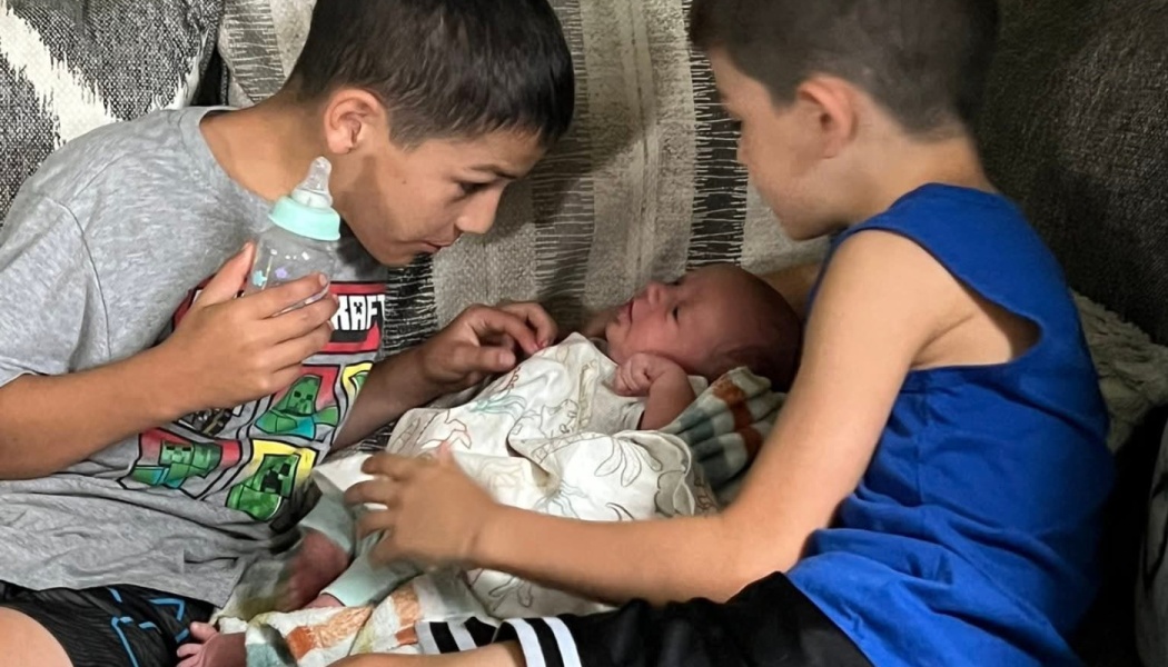 Two young boys lean over a swaddled newborn baby on a couch, with one holding a baby bottle.