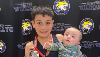 A smiling boy wearing a 2nd-place wrestling medal holds a baby in front of an Onsted Wildcats backdrop.