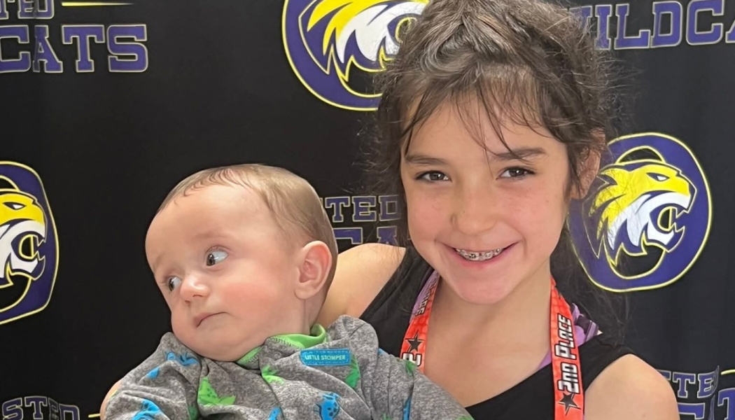 A smiling young girl wearing a 2nd place ribbon holds a baby in front of a Wildcats backdrop.