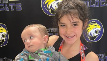A smiling young girl wearing a 2nd place ribbon holds a baby in front of a Wildcats backdrop.