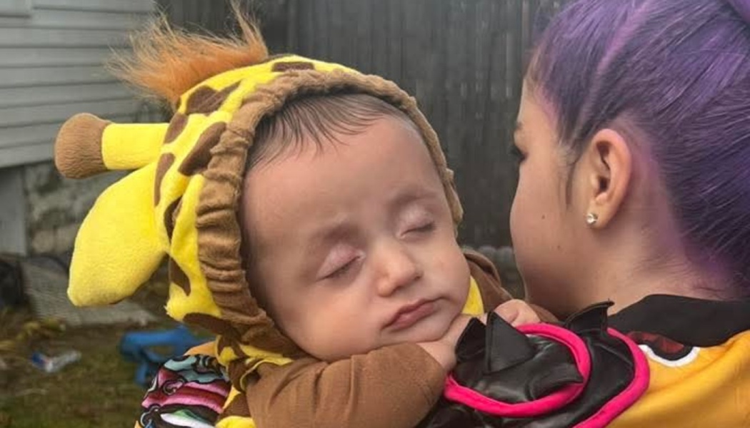 Sleeping baby in a giraffe costume rests peacefully on the shoulder of a person with purple hair.