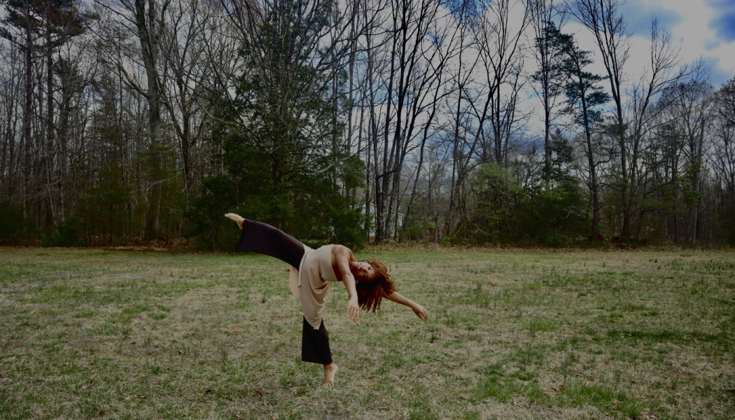 A dancer with red curly hair performs a graceful pose in a grassy field backed by dense woods.