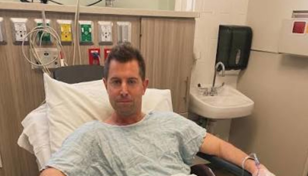 A man in a hospital gown sitting in a hospital bed with medical equipment behind him.