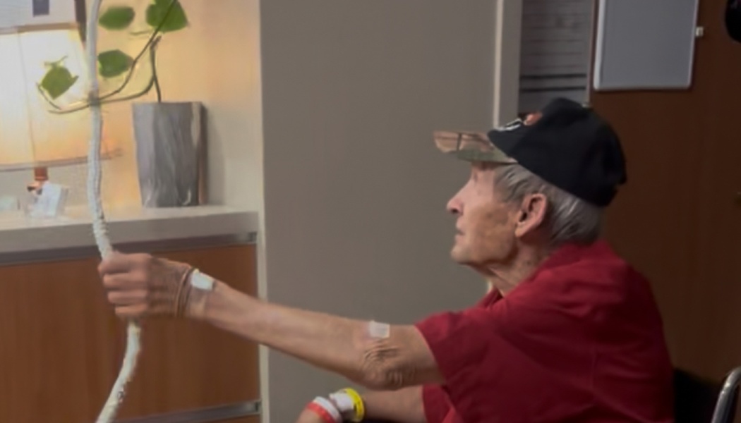 Elderly man in a red shirt and cap wearing hospital bands, reaching out to hold a white wrapped pole.