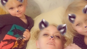 A mother lying down with her two young children, all using playful cat ear digital selfie filters.