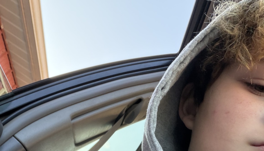 Close-up selfie of a young person wearing a grey hooded sweatshirt inside a car with an open sunroof.
