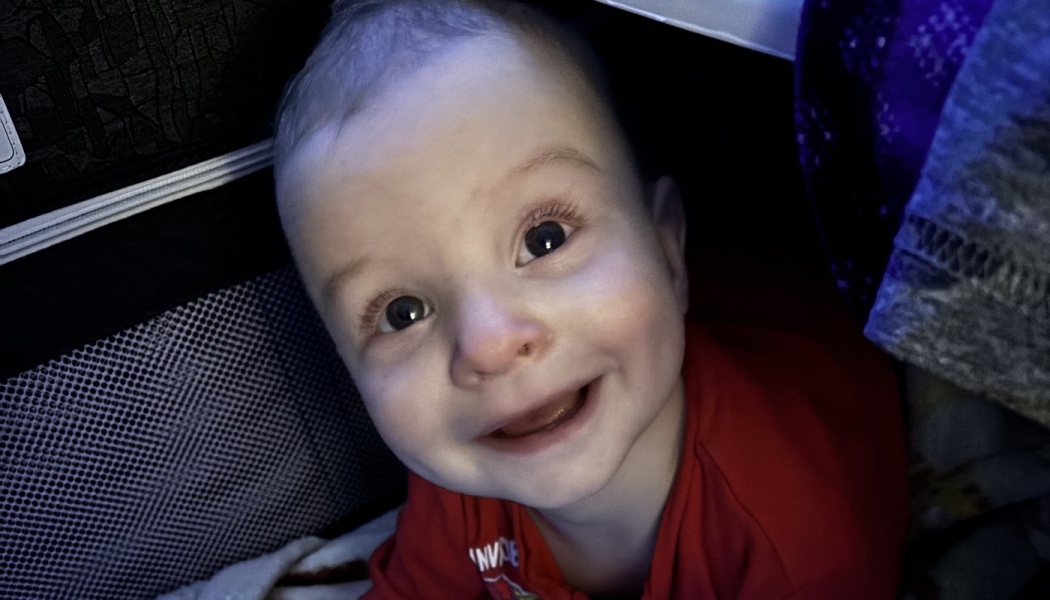 A smiling baby with big, bright eyes looking up at the camera in a red shirt.