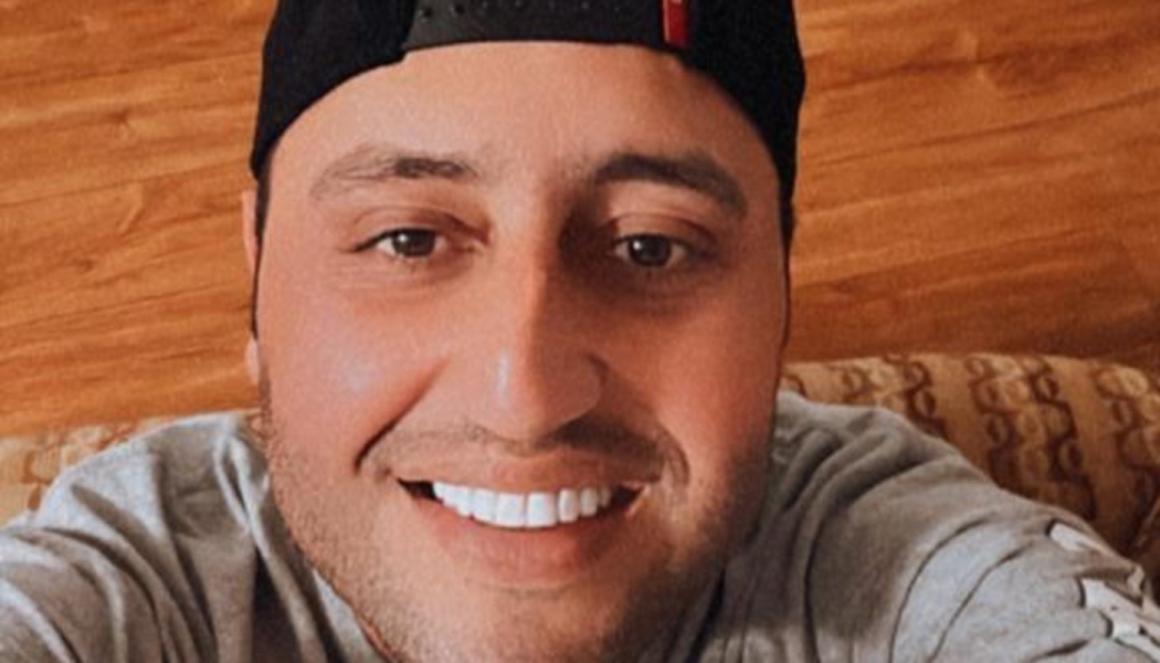 A smiling man wearing a backwards black baseball cap and a grey shirt.
