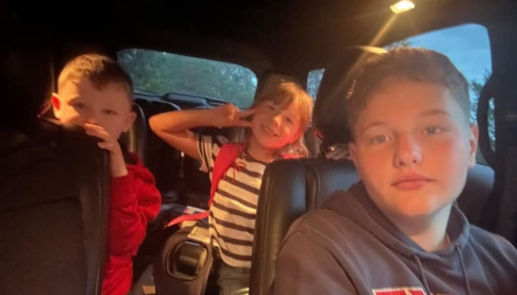 Three children sitting in a car, with a young girl making a peace sign between two boys.