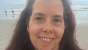 Close-up portrait of a smiling woman with long dark hair, with a sandy beach and ocean in the background.
