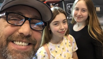 A smiling man in glasses and a baseball cap takes a close-up selfie with two young girls.