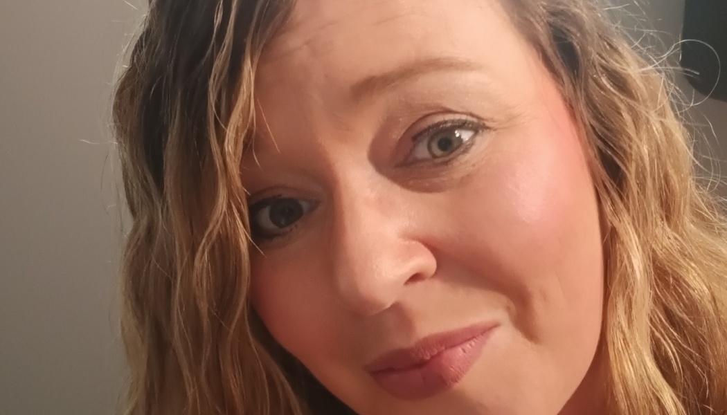 Close-up portrait of a woman with wavy blonde hair and light eyes looking at the camera with a gentle smile.