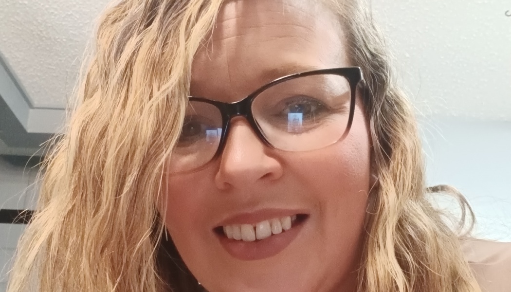 Smiling woman with long blonde wavy hair wearing black-framed glasses.