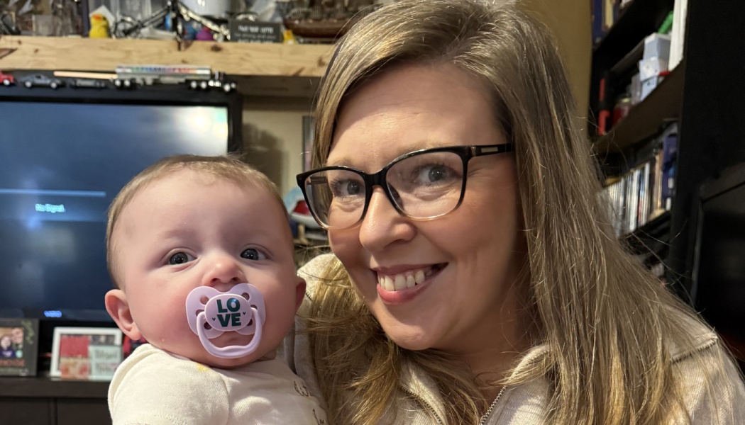 A smiling woman wearing glasses poses next to a baby with a pacifier in its mouth.