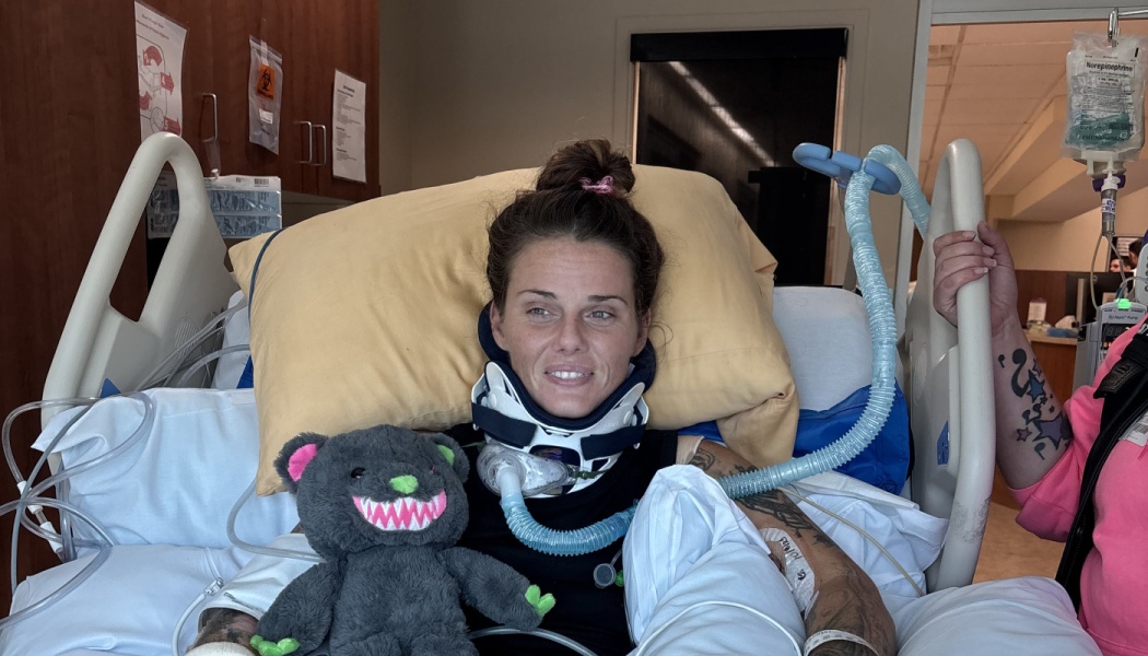 A woman in a hospital bed wearing a neck collar and breathing tube smiles slightly next to a plush stuffed toy.