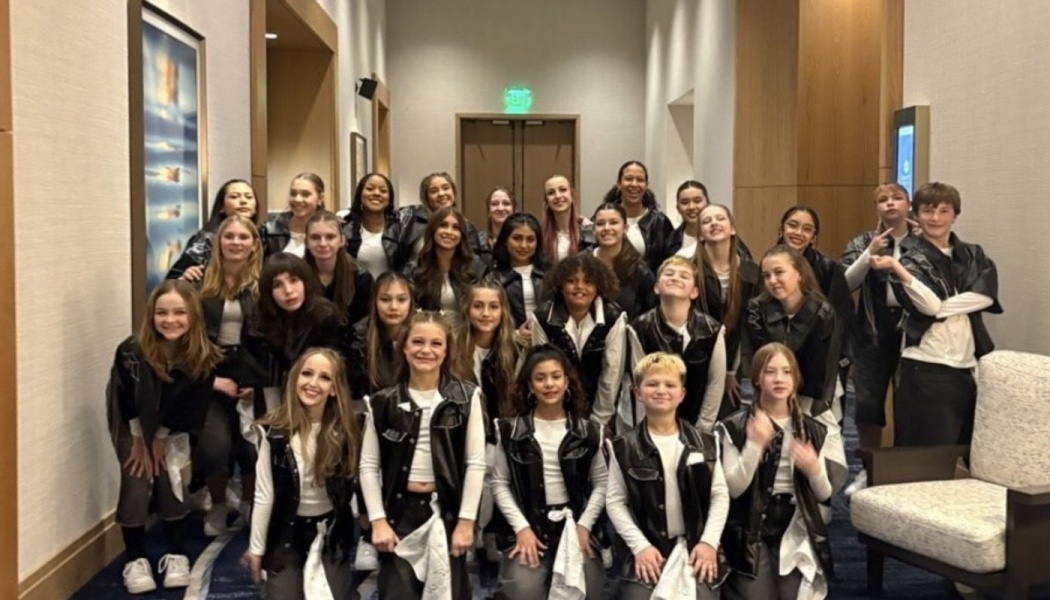 A large youth dance team in matching black leather vests and white tops poses together in a hallway.