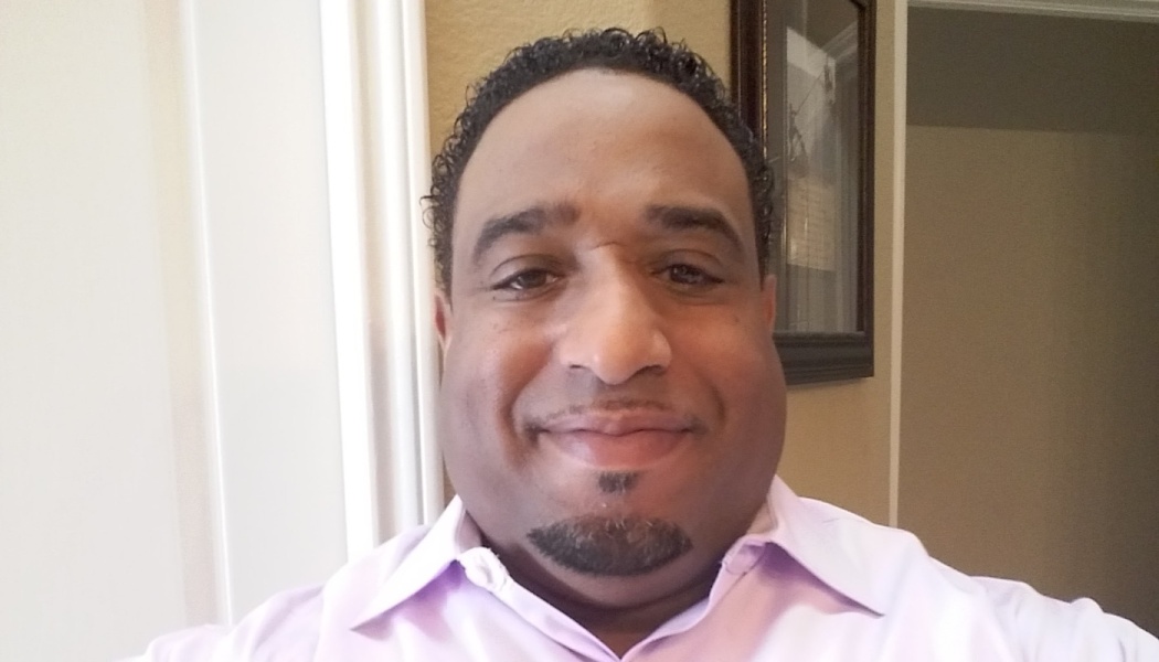 Close-up selfie of a smiling man with a goatee wearing a light pink button-up shirt.
